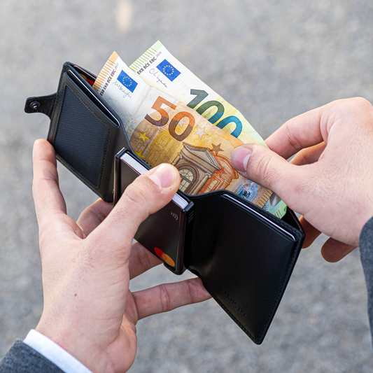 Due manu tengono un portafoglio aperto in mano. La mano destra estrae banconote da 50 e 100€. Sfondo elegante e chiaro.