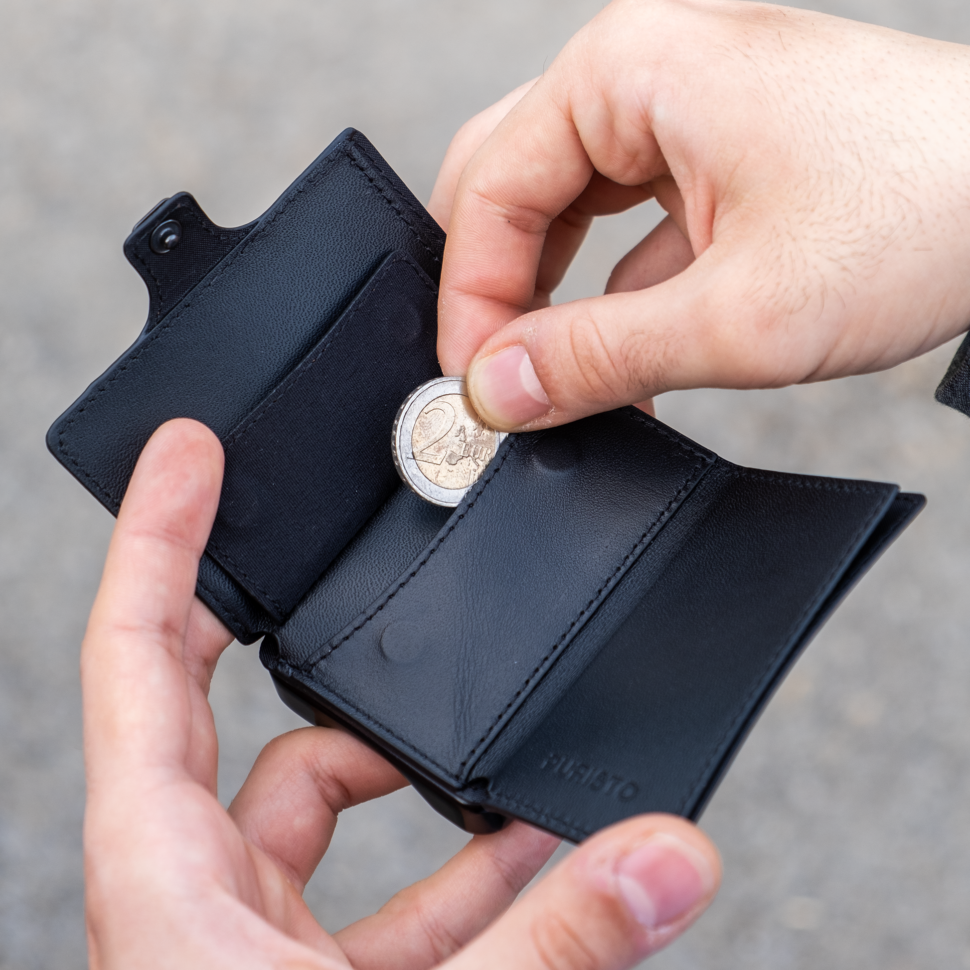 Due mani tengono un portafoglio, la mano destra sfila una moneta da 2 euro. Sfondo chiaro.