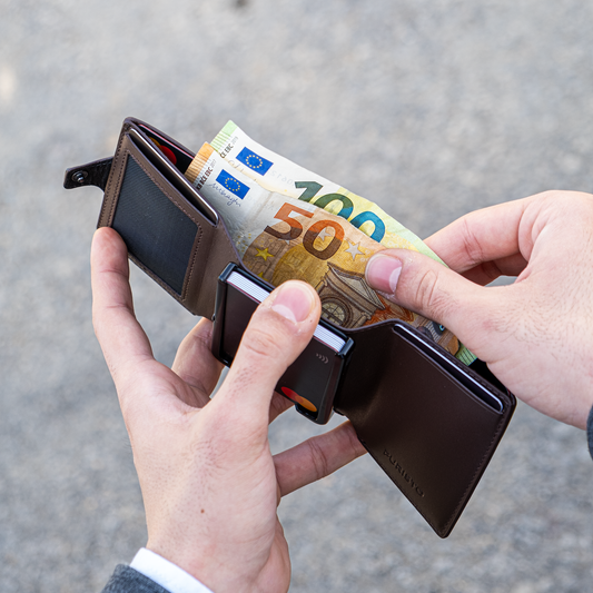 Due manu tengono un portafoglio aperto in mano. La mano destra estrae banconote da 50 e 100€. Sfondo elegante e chiaro.