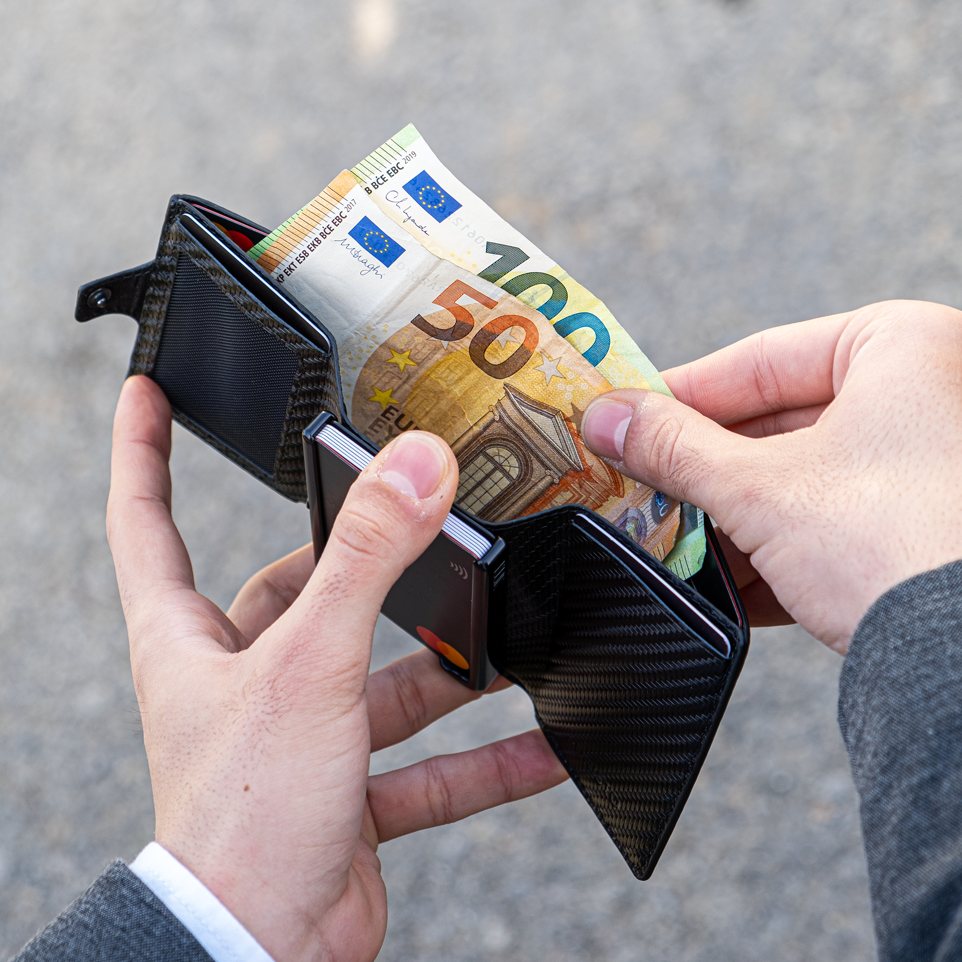 Due manu tengono un portafoglio aperto in mano. La mano destra estrae banconote da 50 e 100€. Sfondo elegante e chiaro.